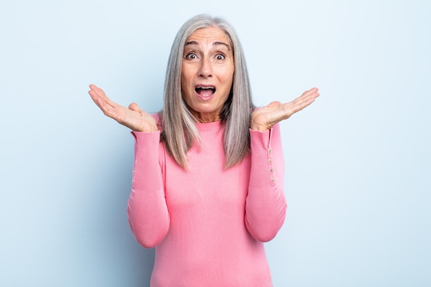 Mujer de cabello gris de mediana edad que parece feliz y emocionada, sorprendida con una sorpresa inesperada con ambas manos abiertas junto a la cara