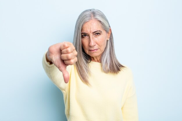 Mujer de cabello gris de mediana edad que se siente enfadada, enojada, molesta, decepcionada o disgustada, mostrando los pulgares hacia abajo con una mirada seria