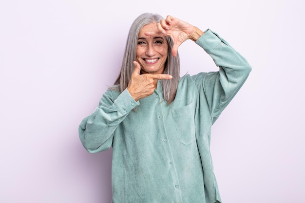 Mujer de cabello gris de mediana edad que se siente feliz, amigable y positiva, sonriendo y haciendo un retrato o marco de fotos con las manos