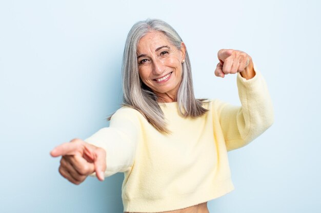 Mujer de cabello gris de mediana edad que se siente feliz y segura, apuntando a la cámara con ambas manos y riendo, eligiéndote