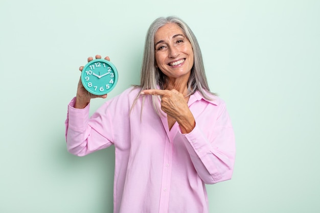 Mujer de cabello gris de mediana edad sonriendo alegremente sintiéndose feliz y señalando el concepto de despertador lateral