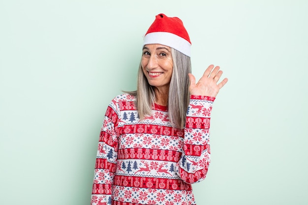 Mujer de cabello gris de mediana edad sonriendo felizmente, saludando con la mano, dándote la bienvenida y saludándote. concepto de navidad