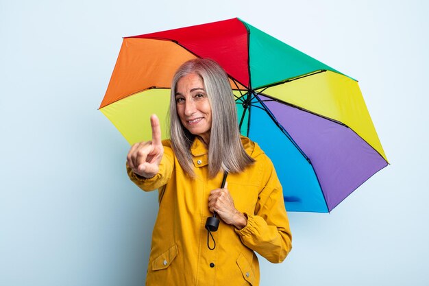 Mujer de cabello gris de mediana edad sonriendo con orgullo y confianza haciendo el número uno. concepto de paraguas y lluvia