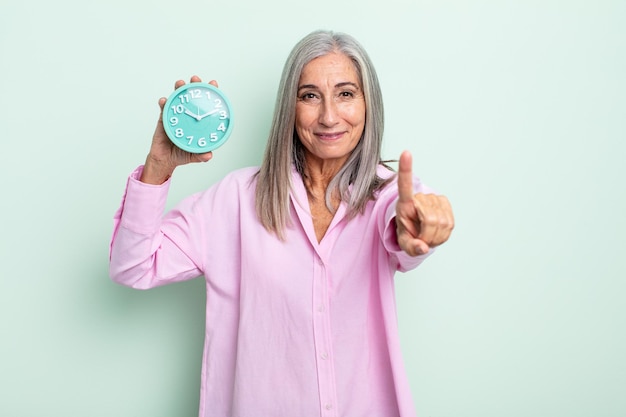 Mujer de cabello gris de mediana edad sonriendo con orgullo y confianza haciendo el número uno. concepto de reloj despertador