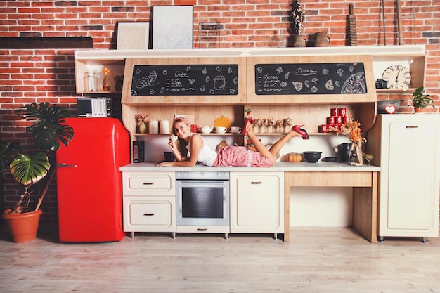 Mujer de cabello largo bastante rubia acostada en la mesa sonriendo y sosteniendo cupcake en la cómoda cocina de loft rojo