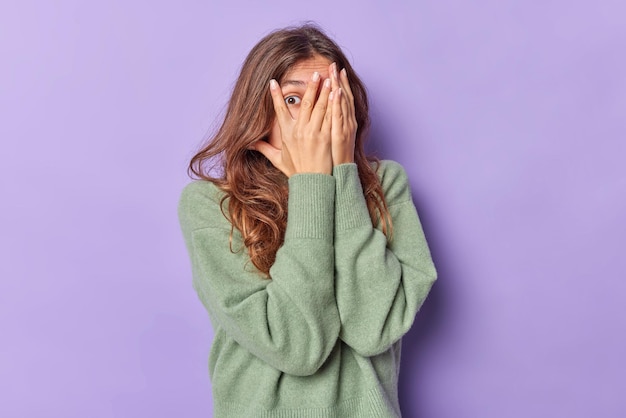 mujer con cabello largo contras la cara con las manos se asoma a través de los dedos temerosa de algo y trata de esconderse viste un jersey casual aislado en morado intrigado por lo que está sucediendo