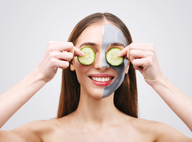 Mujer de cabello largo con máscara de arcilla sosteniendo rodajas de pepino contra sus ojos