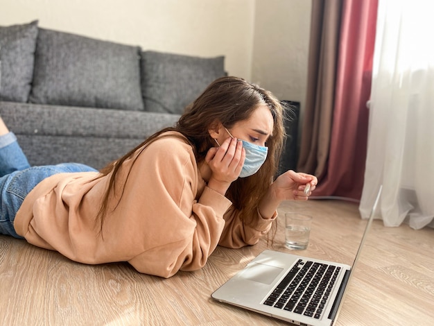Mujer con cabello largo en una máscara médica y un termómetro en sus manos. La mujer tiene signos de fiebre a alta temperatura. Coronavirus pandémico