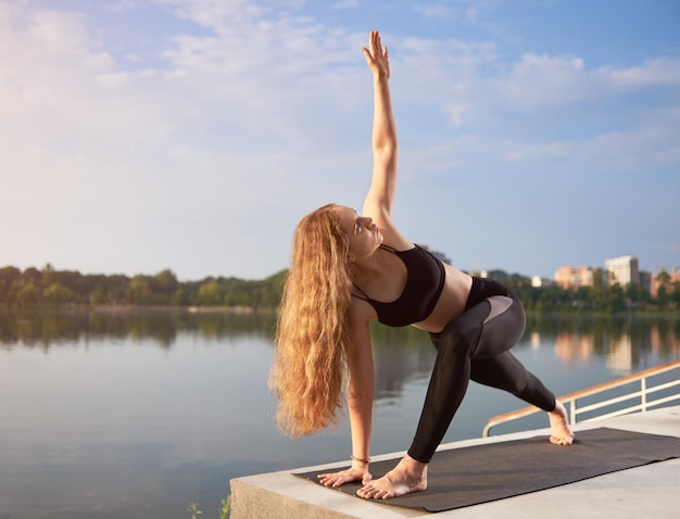 Mujer con cabello largo en ropa deportiva negra haciendo yoga en una alfombra de yoga cerca del lago por la mañana