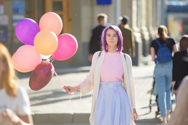 Foto mujer con cabello morado de pie en la calle con un montón de globos aerostáticos