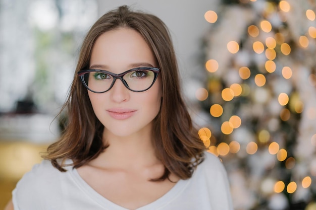 Mujer de cabello oscuro con gafas expresión seria está junto al árbol de Año Nuevo en el interior mirada atractiva