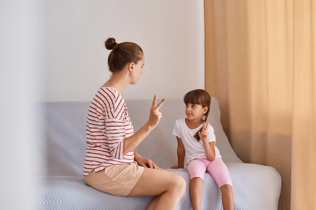 Mujer de cabello oscuro con patólogo del habla de moño que tiene una lección con una niña, entrenando la pronunciación de los sonidos y la articulación, gente sentada en un cómodo sofá junto a la ventana.