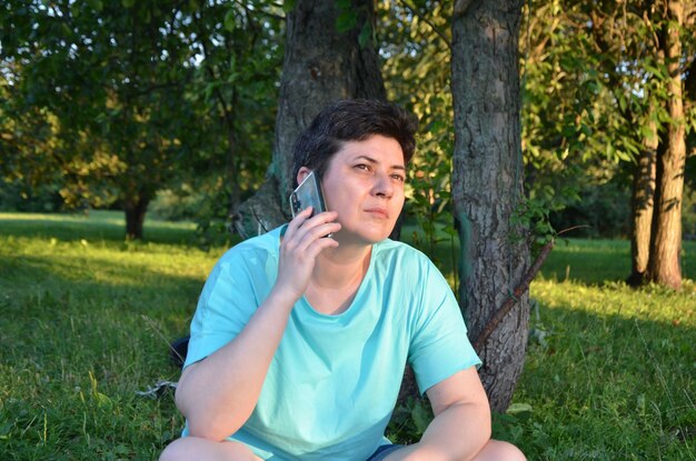 Mujer de cabello oscuro con el pelo corto con una camiseta azul y pantalones cortos se sienta en la hierba en el parque, hablando por un teléfono celular. mujer durante una conversación telefónica.