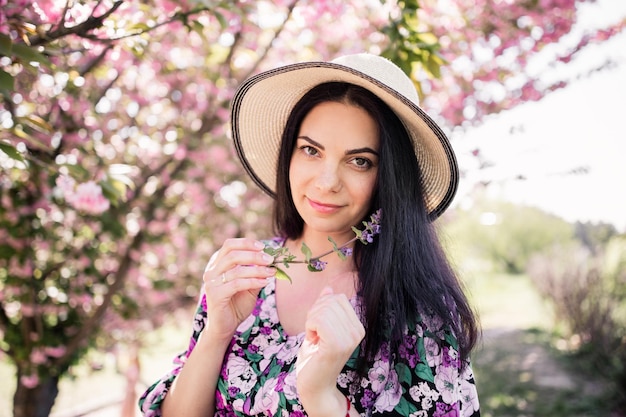 Mujer con cabello oscuro en un sombrero de paja cerca de flores de cerezo en primavera