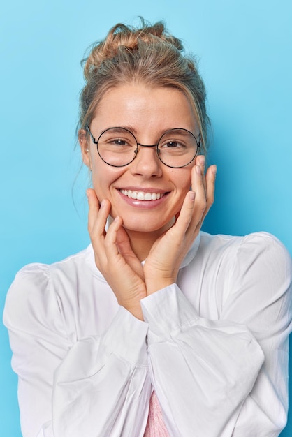 Mujer con cabello peinado, sonrisas positivas en la cara, toca las mejillas, usa gafas redondas y camisa blanca aislada en azul, llega a la oficina para una entrevista de trabajo