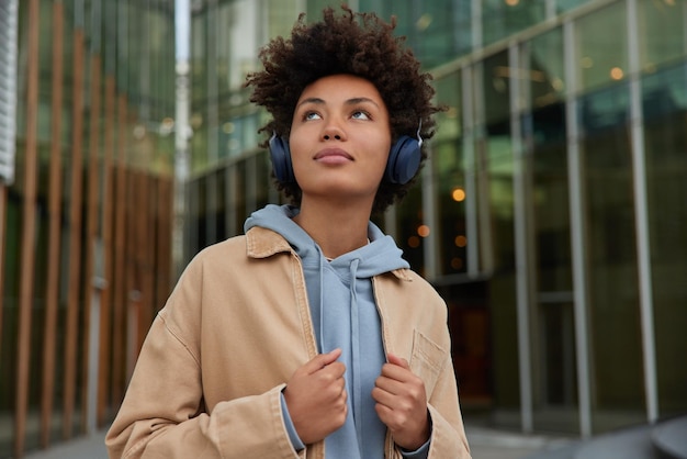 mujer con cabello rizado enfocado arriba escucha música favorita en auriculares usa sudadera con capucha casual y poses de chaqueta contra la entrada de vidrio al centro comercial disfruta de un buen sonido piensa en algo