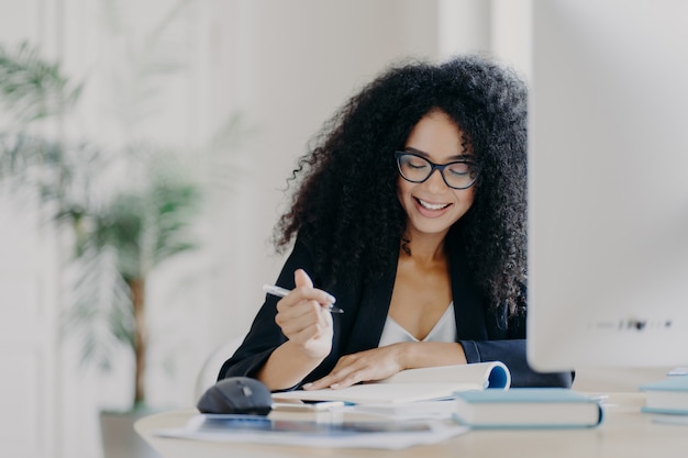 La mujer de cabello rizado escribe alguna información, sostiene la pluma, sonríe y usa lentes ópticos