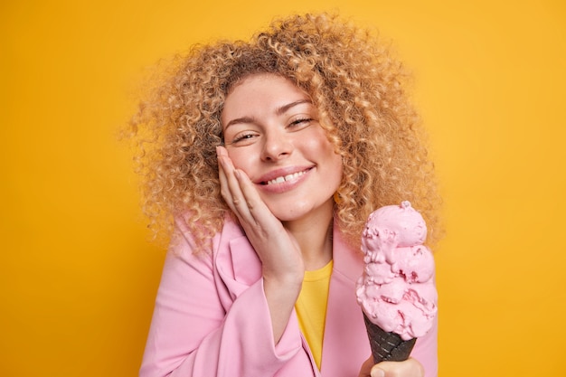 Mujer con cabello rizado mantiene la mano en la mejilla sonríe suavemente sostiene sabroso helado siente la tentación de comer dulce postre frío expresa felicidad