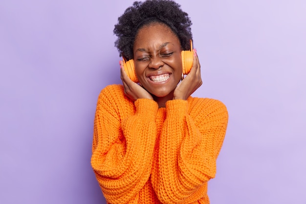 Mujer con cabello rizado mantiene las manos en los auriculares disfruta de un buen sonido agradable melodía vestida con un jersey de punto naranja aislado en violeta