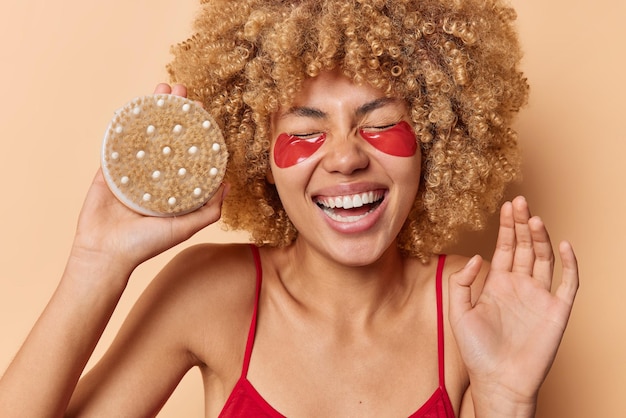 Una mujer de cabello rizado muy contenta se ríe felizmente sostiene el cepillo corporal mantiene las sonrisas levantadas con la palma de la mano y aplica parches de belleza de hidrogel debajo de los ojos para eliminar las arrugas aisladas sobre el fondo marrón