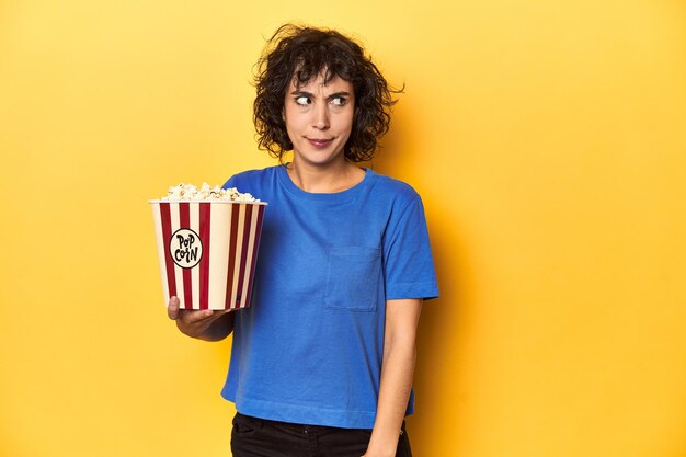 Mujer de cabello rizado con palomitas de maíz para el estudio de cine confundida se siente dudosa e insegura