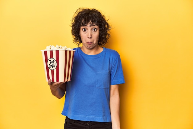 Mujer de cabello rizado con palomitas de maíz para el estudio de cine se encoge de hombros y los ojos abiertos confundidos