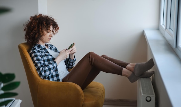 Mujer de cabello rizado con ropa cómoda escribiendo en la pantalla del teléfono inteligente sentada en una silla de co...