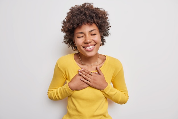 mujer con cabello rizado sonrisa sincera en la cara presiona las manos en el pecho se siente agradecida por la ayuda cierra los ojos viste un jersey amarillo casual posa en blanco halagado de algo reconfortante