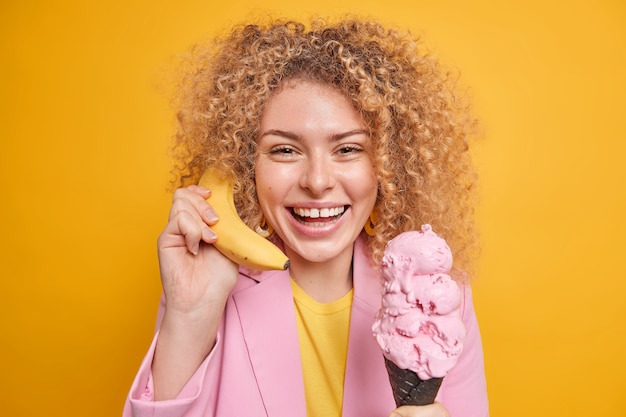 Mujer con cabello rizado sostiene un delicioso plátano cerca de la oreja llama a un amigo come un delicioso helado tiene un estado de ánimo optimista y sonríe ampliamente