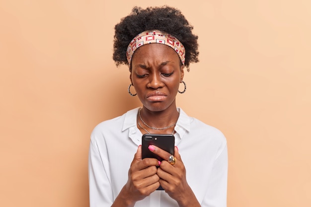 mujer con cabello rizado usa tipos de teléfonos móviles modernos mensajes de texto usa diadema camisa blanca molesta por no tener suficiente dinero en la cuenta bancaria en la aplicación de teléfono inteligente