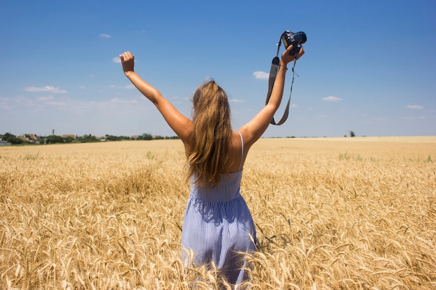 mujer con cabello rubio natural de pie en un campo de trigo con una cámara
