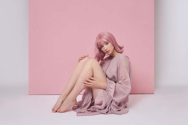 Mujer con cabello teñido de rosa con un vestido largo está sentada en el suelo. Retrato de una niña con coloración del cabello en la pared rosa. Peinado y peinado perfectos.