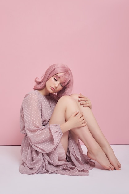 Mujer con cabello teñido de rosa con un vestido largo está sentada en el suelo. Retrato de una niña con la coloración del cabello en la pared rosa