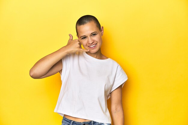 Mujer de cabeza afeitada con camiseta blanca y fondo amarillo que muestra un gesto de llamada de teléfono móvil con los dedos