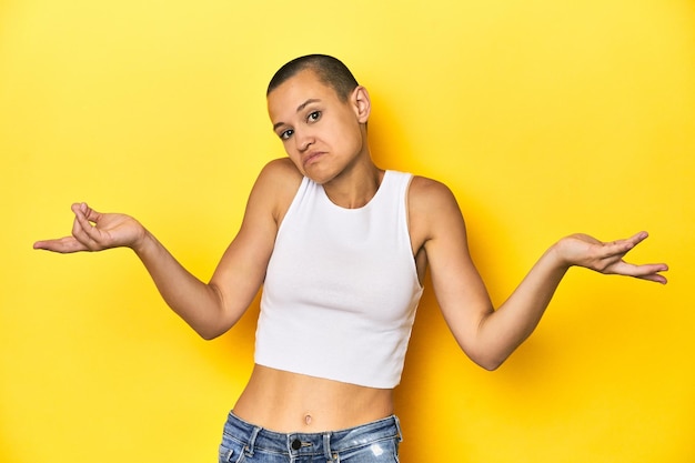 Mujer de cabeza afeitada con camiseta blanca con telón amarillo en el fondo dudando y encogiéndose de hombros