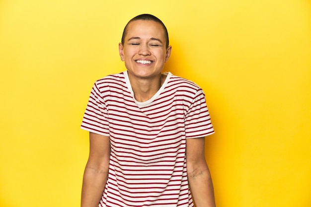 Una mujer con la cabeza afeitada y una camiseta a rayas rojas con fondo amarillo se ríe y cierra los ojos, se siente relajada y feliz