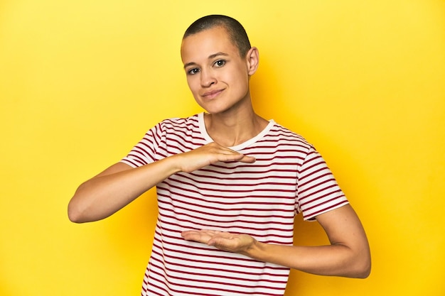 Mujer de cabeza afeitada en camiseta de rayas rojas con telón de fondo amarillo sosteniendo algo con ambas manos