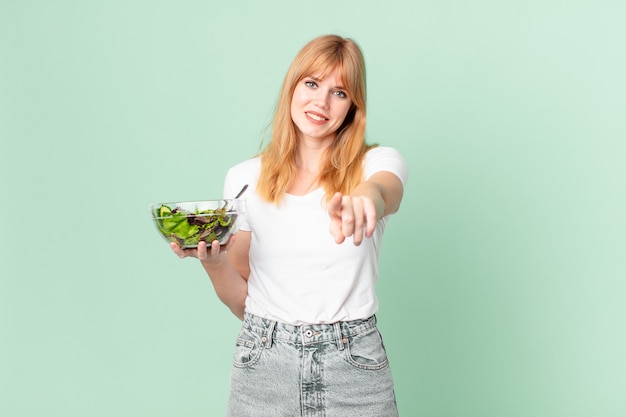 Mujer de cabeza bastante roja apuntando a la cámara eligiéndote y sosteniendo una ensalada. concepto de dieta