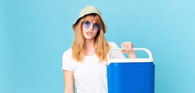 Mujer de cabeza bastante roja que parece perpleja y confundida y que sostiene un refrigerador portátil. concepto de verano