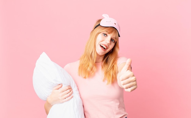 Mujer de cabeza bastante roja que se siente orgullosa, sonriendo positivamente con los pulgares hacia arriba. en pijama y sosteniendo una almohada