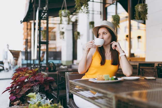 Mujer en el café de la cafetería de la calle al aire libre sentado en la mesa con sombrero amarillo con taza de capuchino, pastel, relajarse en el restaurante durante el tiempo libre