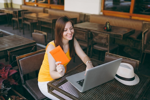Mujer en el café de la cafetería de la calle al aire libre sentado en la mesa trabajando en una computadora portátil moderna, sostiene un pasaporte en la mano, reserva un boleto de avión en línea