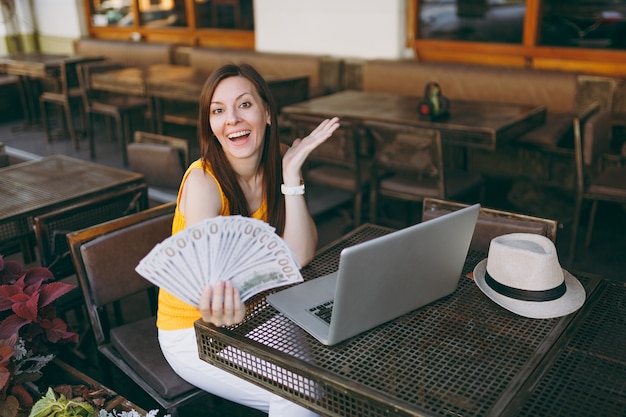 Mujer en el café de la cafetería de la calle al aire libre sentado con un moderno ordenador portátil, tiene en la mano un montón de billetes de dólares