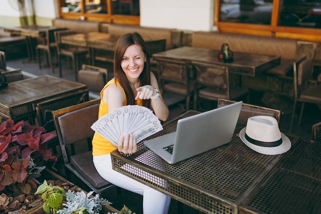 Mujer en el café de la cafetería de la calle al aire libre sentado con un moderno ordenador portátil, tiene en la mano un montón de billetes de dólares