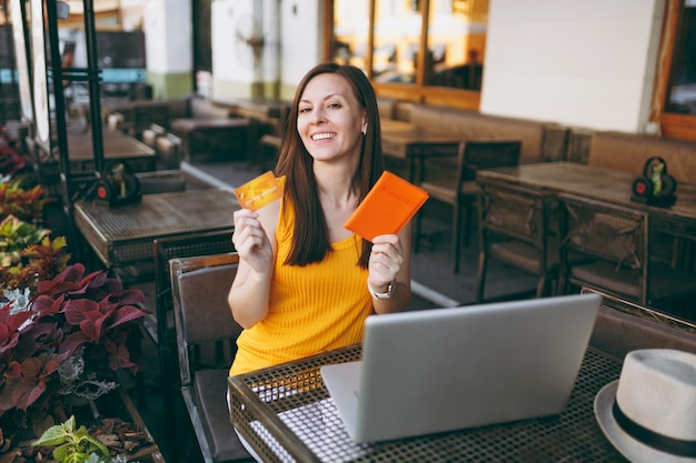 Mujer en el café de la calle al aire libre sentado a la mesa con un moderno ordenador portátil, tiene en la mano la tarjeta de crédito bancaria y el pasaporte