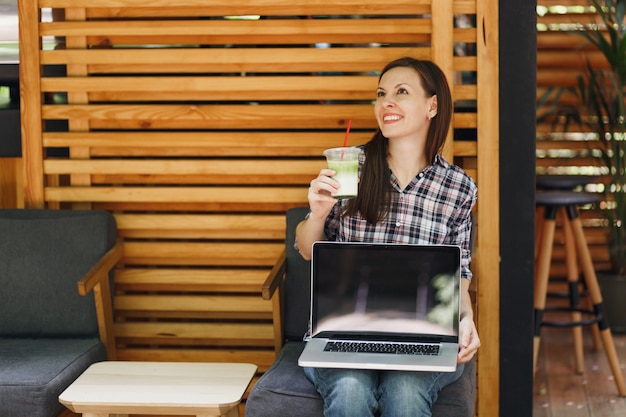 Mujer en el café de madera de la cafetería de la calle al aire libre sentado en ropa casual, sostenga la computadora portátil con pantalla vacía en blanco, relajándose en el tiempo libre. Oficina móvil