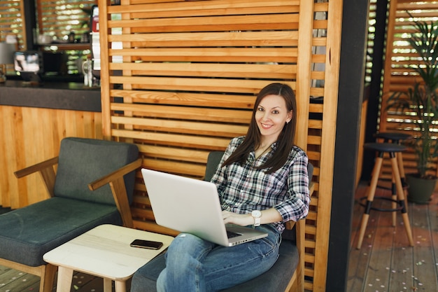 Mujer en el café de madera de la cafetería del verano de la calle al aire libre que se sienta en ropa casual, trabajando en la computadora portátil moderna de la pc