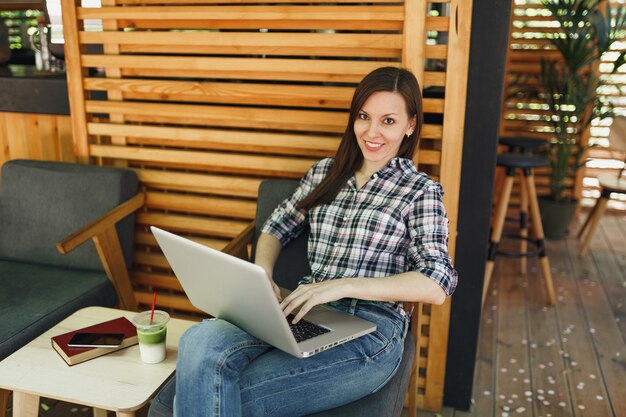 Mujer en el café de madera de la cafetería del verano de la calle al aire libre que se sienta en ropa casual, trabajando en la computadora portátil moderna de la pc