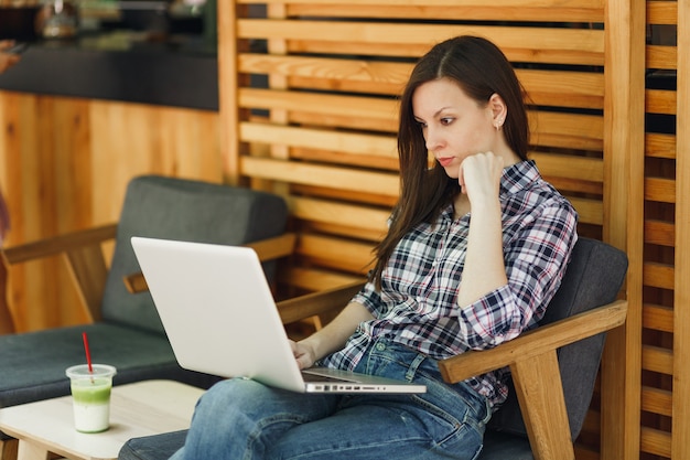 Mujer en el café de madera de la cafetería de verano de la calle al aire libre sentado en ropa casual, trabajando en la computadora portátil moderna, relajándose durante el tiempo libre. Oficina móvil