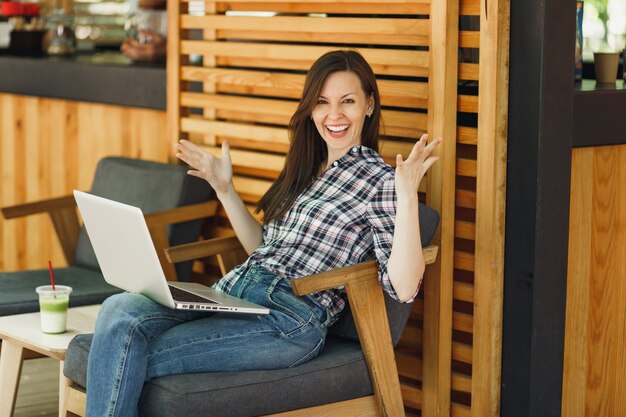 Foto mujer en el café de madera de la cafetería de verano de la calle al aire libre sentado en ropa casual, trabajando en la computadora portátil moderna, relajándose durante el tiempo libre. oficina móvil